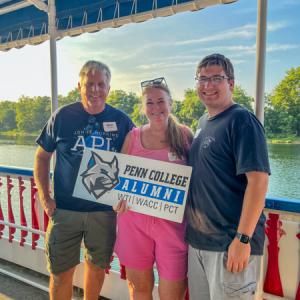 A group of three holds a Penn College Alumni sign. Behind them are a red and blue railing, water, trees and blue skies.