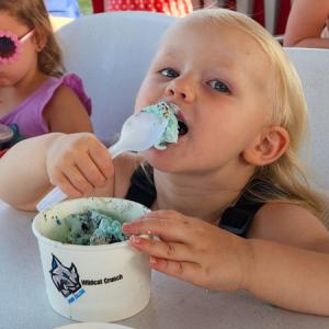 A small child lifts a spoonful of light green ice cream with chocolate flecks to her mouth.
