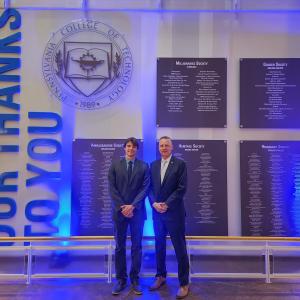 Two men in business suits stand in front of a wall that shows the Pennsylvania College of Technology seal and several plaques that list names.