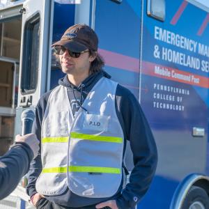 A student in a reflective vest and baseball hat speaks into a microphone held by another person who is not shown. Behind the student is a vehicle that reads: "Emergency Management & Homeland Security, Mobile Command Post, Pennsylvania College of Technology."