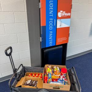 A wagon full of food sits in front of a mini cupboard, which is a cabinet and mini refrigerator, in a Hager Lifelong Education Center hallway.