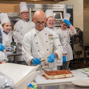 While students in chefs uniforms gather behind him, a chef dusts cocoa powder onto a cake.