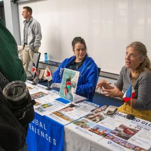 Two women sit at a table covered in Global Experience pamphlets. One is talking to a person who is partially obscured in the foreground.