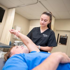 A clinician assists a person lying on a treatment table by guiding their arm through a range‑of‑motion exercise in a physical therapy setting.
