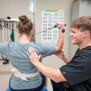 Physical therapy session where a person sits on an exercise ball pulling resistance bands while a clinician provides guidance and support at their back.