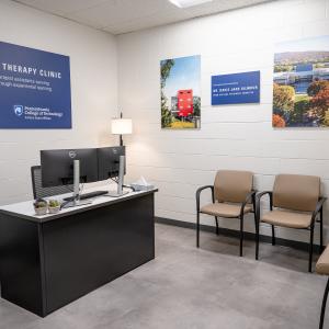 Reception-style room for a physical therapy clinic with a desk, chairs for visitors, and wall art featuring campus and clinic imagery.