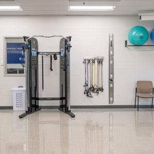 Physical therapy room featuring a cable machine, resistance bands on the wall, therapy balls on a shelf, and a chair positioned nearby