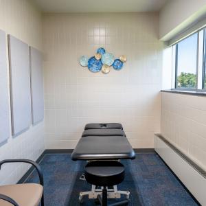 Small treatment room with a therapy table, a stool, wall décor, and a window letting in natural light.