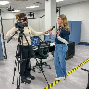 A student talks with a reporter who is holding a microphone and operating a TV camera.