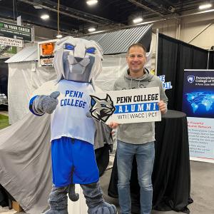 Person stands beside a cut-out of the Penn College Wildcat mascot, holding a Penn College Alumni sign at a booth.