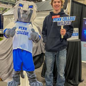 Person stands beside a cut-out of the Penn College Wildcat mascot, holding a Future Penn College Wildcat sign at a booth.