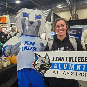Person stands beside a cut-out of the Penn College Wildcat mascot, holding a Penn College Alumni sign at a booth.