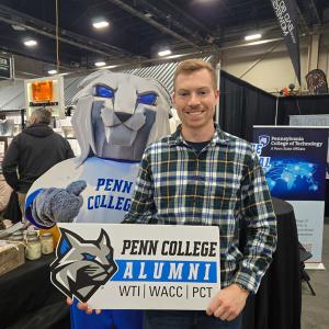Person stands beside a cut-out of the Penn College Wildcat mascot, holding a Penn College Alumni sign at a booth.
