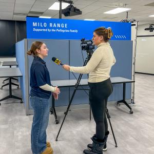 A student talks with a reporter who is holding a microphone and operating a TV camera.