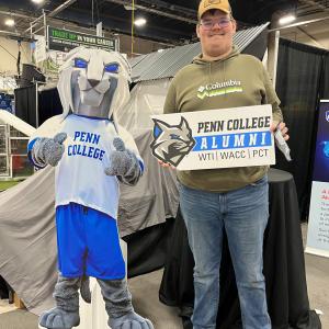 Person stands beside a cut-out of the Penn College Wildcat mascot, holding a Penn College Alumni sign at a booth.