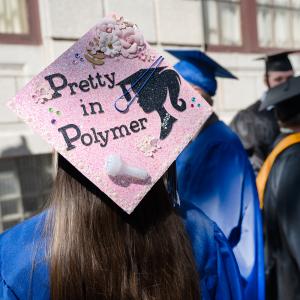 A graduation cap is decorated in pink with silk flowers, a vial of plastic pellets, and the words "Pretty in Polymer."