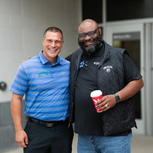 Two men smile widely while standing outside the door of a Penn College building. One is holding a disposable coffee cup.