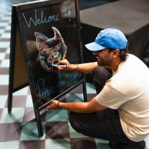 A student uses markers to draw a Wildcat on a black dry-erase easel that has the handwritten words "Welcome to Penn College."