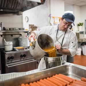 In a commercial kitchen at Little League International, a man in a chef uniform pours soup from a large pot into a stainless steel container.