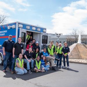 Penn College emergency management and homeland security students and faculty pose outside of a vehicle that has the words “Emergency Management & Homeland Security Mobile Command Post, Pennsylvania College of Technology.” Penn College’s main entrance fountains and Madigan Library are visible in the background. 
