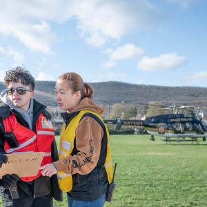 Penn College emergency management and homeland security students wearing brightly colored safety vests talk while looking over a clipboard at the program’s annual Rotor Fest. A state police helicopter is visible on a college lawn behind them. 