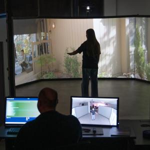 A man sits behind a set of computers while a student stands inside a circle of projection screens. The student interacts with a person projected onto the screens.