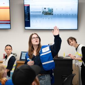 A student stands in the middle of a classroom pointing toward a monitor on the wall. All students are wearing different-colored vests.