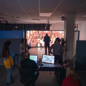A state legislator conducts a simulated press conference inside a circle of large projection screens that show a group of reporters. Several people watch from outside the circle. A person sits behind a computer to control the simulation.