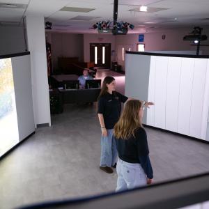 Students stand inside a circle of large projection screens and interact with the characters projected onto the screens. A person sits behind a computer just outside the circle to control the simulation.