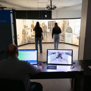 Students stand inside a circle of large projection screens and interact with the characters projected onto the screens. A person sits behind a computer just outside the circle to control the simulation.