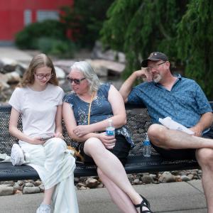 A teen and two adults sit on a bench outside Penn College's Madigan Library. The teen is sitting on the left holding and looking at a phone, and a woman sitting next to her is looking over the teen's shoulder at the phone.