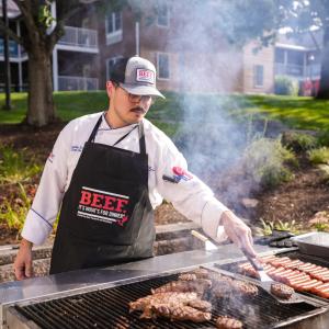 A man in a chef uniform and "Beef: It's what's for dinner" apron flips hamburgers on a large grill. Also on the grill are steaks and hot dogs.