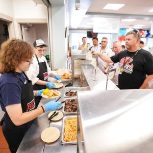 Two people standing behind a cafeteria service line add food to plates as coaches and players wait on the other side.