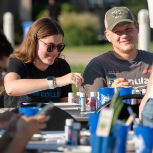 Students sit at an outdoor table painting canvases.