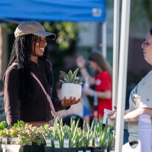 A student holds a plant in a decorative container and speaks with a woman behind a table of several types of succulent plants.