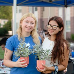 Two students pose holding plants in coral-colored pots.