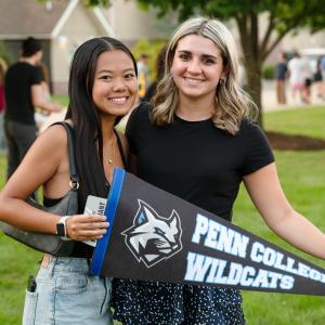 Two studnets stand outdoors holding a Penn College Wildcats pennant.