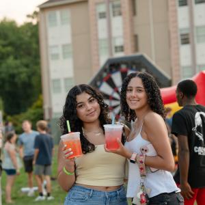 Two students pose holding slushy drinks. Other students and inflatable games are visible in the background, as is a Penn College residence hall.
