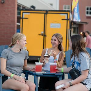 Three students sit at an outdoor table smiling and chatting. A food truck, students, and Penn College's Dauphin Hall are behind them.