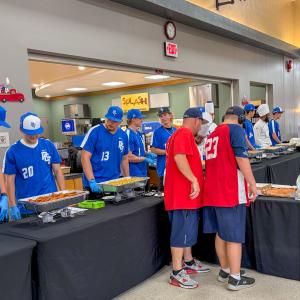 Students in blue Penn College baseball jerseys stand behind tables to serve food to people in red baseball jerseys. (Photo by Danielle 