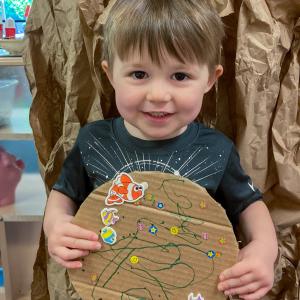 A small child holds a cardboard circle on which he has colore and placed stickers.