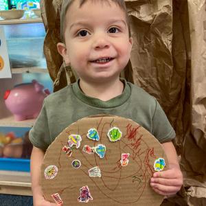 A small child holds a cardboard circle on which he has colore and placed stickers.