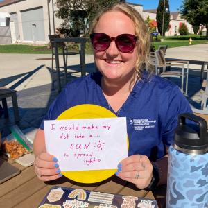 A physician assistant student wearing blue scrubs holds up a yellow dot and a paper with the handwritten words: "I would make my dot into a sun to spread light."