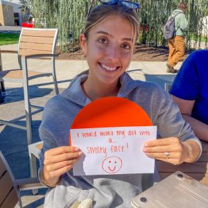 A physician assistant student wearing blue scrubs and a sweatshirt holds up an orange dot and a paper with the handwritten words: "I would make my dot into a smiley face."