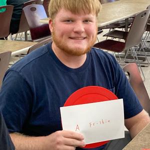 A student holds up a red circle and a piece of paper on which he as written the word "Frisbie."