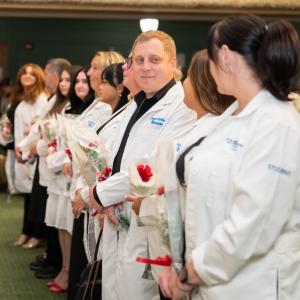 Graduating students look at one another with smiles as they stand in the front row of Penn College's Klump Academic Center Auditorium. They are wearing white lab coats.