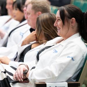 Graduating students wearing white lab coats laugh at a speakers comments while they sit in the front row of Penn College's Klump Academic Center Auditorium.