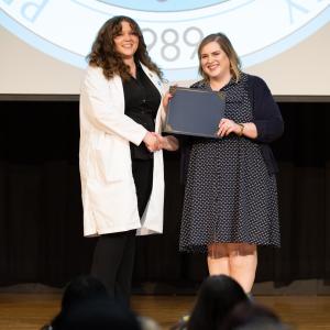 Two women stand on a stage shaking hands and holding a diploma folder. One is wearing a white lab coat.