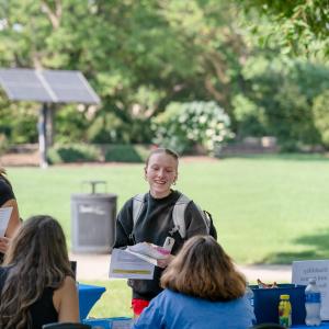 A student holding a handful of brochures and giveaways smiles while talking to a woman seated at a table.