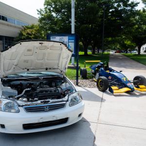 A white Honda with its hood open and a blue formula-style vehicle are displayed along a Penn College sidewalk.